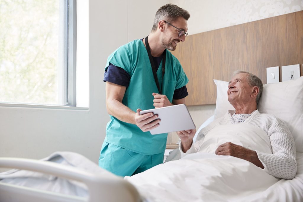Surgeon With Digital Tablet Visiting Senior Male Patient In Hospital Bed In Geriatric Unit